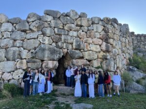 Group of people, mostly young adults, posing in front of a tall stacked stone wall outdoors under a clear blue sky.