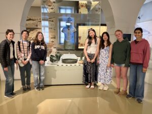 Eight youths pose in a museum gallery in front of glass display cases with ancient artifacts and pottery behind them, smiling at the camera.