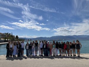 Group of people posing together on a waterfront promenade with mountains in the distance, blue sky above.