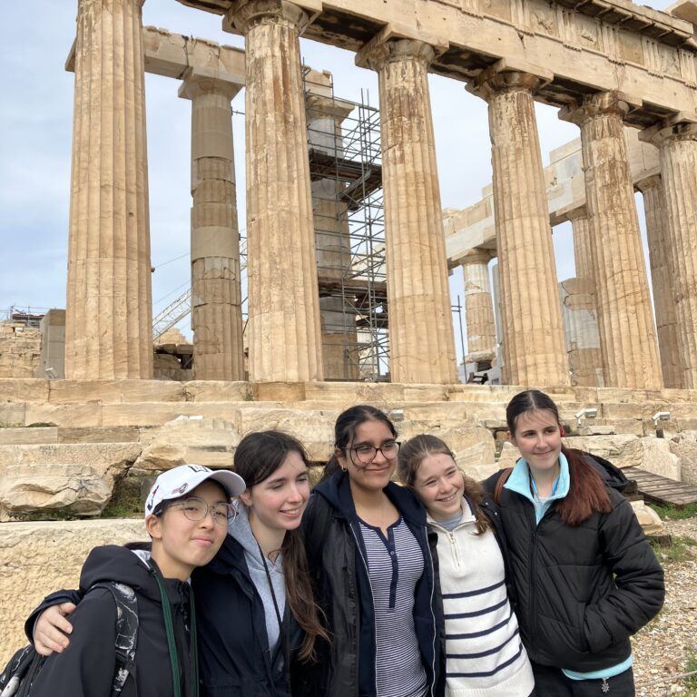 Five students pose together in front of ancient stone temple columns, with scaffolding visible in the background.