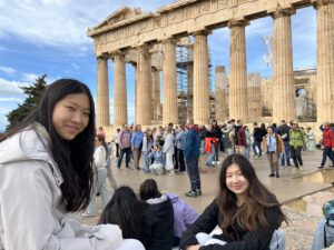Group of tourists posing near ancient Greek ruins with tall stone columns and scaffolding in the background under a blue sky.