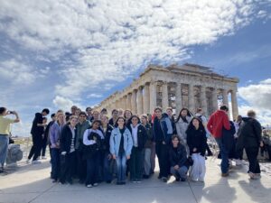 Group of students posing for a photo in front of the Parthenon-like temple under construction scaffolding, clear blue sky above.