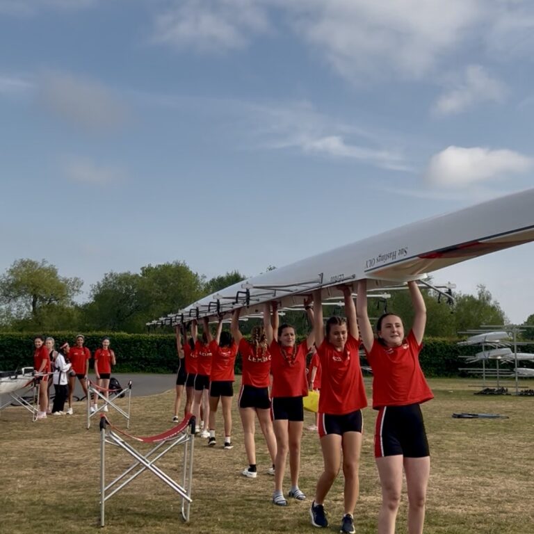 Rowing team in red shirts lift a long white racing shell overhead in a grassy field under a blue sky.