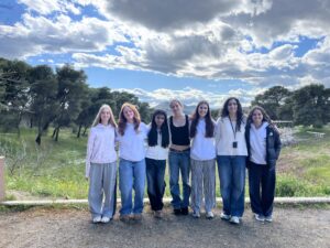 Group of young people standing together outdoors, smiling, in a grassy area with trees and a cloudy blue sky behind them.