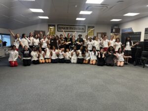 Group of students in white shirts posing and waving in a classroom with a Keystone Productions banner on the wall behind them.