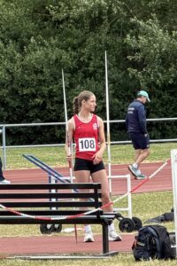 Athlete in red top with bib 108 holding a pole vault pole on a track field near a bench and barriers.