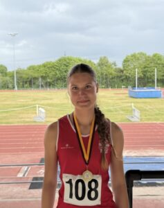 Female athlete with bib 108 and a gold medal on a ribbon, standing on a track with a grassy field in the background.