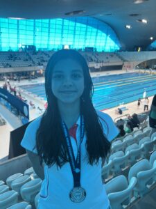 Girl at an indoor swimming pool arena wearing a lanyard and medal, smiling at the camera with blue seating in the background.