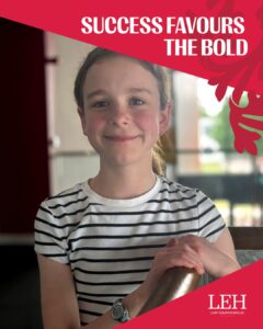 Smiling girl in a striped shirt indoors, with a pink diagonal banner reading 'SUCCESS FAVOURS THE BOLD' and a LEH logo in the corner.