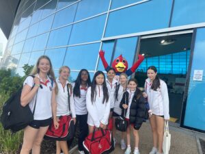 Group of smiling schoolgirls in white uniforms with red trim, carrying bags, standing outside a blue glass building near a red mascot in a costume.