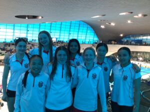 Group of smiling young athletes in matching white jackets posing together inside a modern indoor swimming arena.