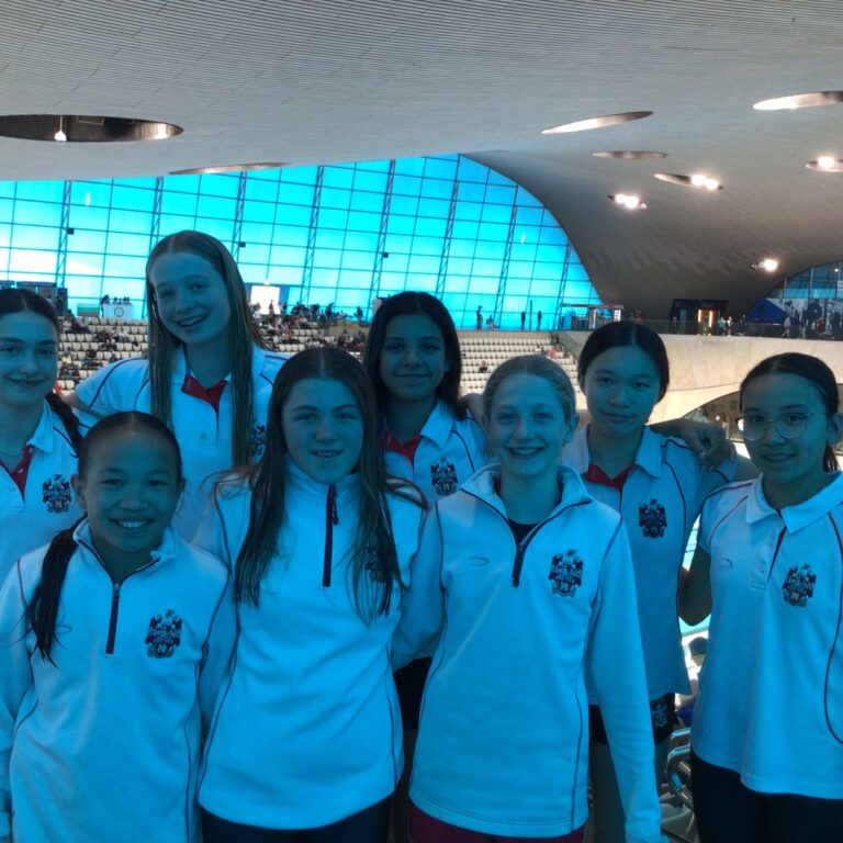 Group of smiling young athletes in matching white jackets posing together inside a modern indoor swimming arena.