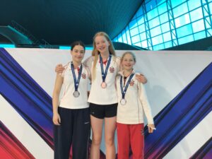 Three young female athletes on a podium wearing white shirts with medals around their necks, posing for a photo with a colorful backdrop behind them.