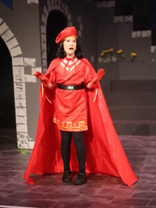 Performer in a bright red cape and hat on stage, making a surprised expression with gloved hands raised.
