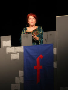 Actress in a green velvet costume reads a small book on stage, standing behind gray panels and a blue banner with a red symbol.