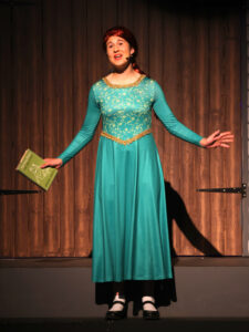 Young girl on stage in a turquoise dress with gold trim, holding a green book and wearing a headset microphone, arms extended.