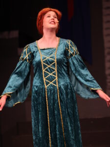 Woman on stage in a teal velvet Renaissance dress with gold trim, arms outstretched, singing into a headset microphone.