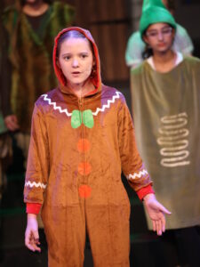 Young girl on stage wearing a brown costume with orange buttons and a red hood, speaking into a headset microphone.