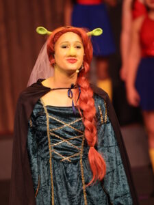 Person in a Shrek-inspired costume on stage: green headband ears, red braided wig, teal velvet gown with gold trim, cape, and veil.