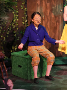 Child wearing tiger-face makeup and round glasses, in a blue blazer and plaid pants, seated on a green stage prop during a performance.