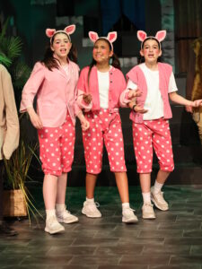Three young girls in pink pig-costume outfits perform on a stage, wearing pig ears and white sneakers.