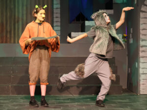 Theater scene with two performers: one in an orange poncho and plaid pants holding a prop, the other in a gray fur wolf costume mid-leap on stage.