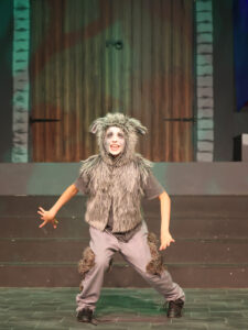 Child in a fluffy gray wolf costume with ears performing on a theater stage, arms outstretched and smiling