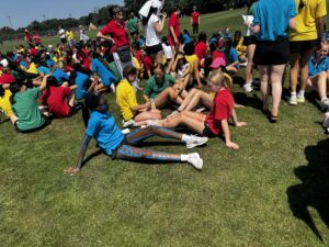Large group of students in colorful shirts gathered on a grassy sports field during an outdoor event on a sunny day.