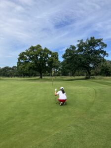 Golfer kneeling on putting green, preparing for a putt near the hole with a flagstick and blue sky above.