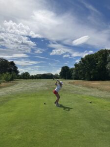 Person in white hat and red shorts mid-swing on a golf tee box, preparing to drive the ball on a sunny course with green fairway and trees in the distance.