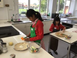 Two girls in red uniforms and green aprons chopping vegetables at a bright school kitchen counter in a cooking class setting.