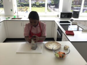 Person wearing a red apron grates cheese on a box grater at a white kitchen island, with bowls of chopped vegetables and dough nearby and a bright window view behind.