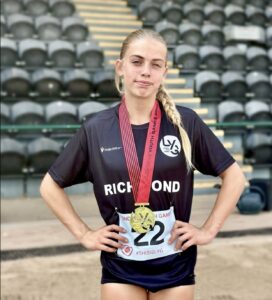 Female athlete in a black Richmond shirt with a gold medal around her neck, standing in a stadium with rows of seats behind her.