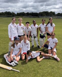 A youth girls' cricket team posing on a sunny field in matching white uniforms with red trim, some wearing pads and helmets, and a bat lying on the grass in front.