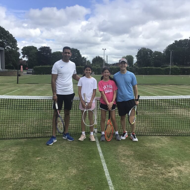 Four people stand together on a grass tennis court with rackets and a net, smiling at the camera under a cloudy sky.
