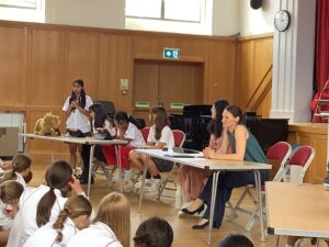 Girl in white shirt speaks into a microphone at a seated table of students; a teddy bear sits on the table beside her in a school hall setting.