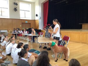 Student presenter speaks into a microphone at a desk in a school assembly, addressing seated classmates in white uniforms; a pony costume is in the foreground.