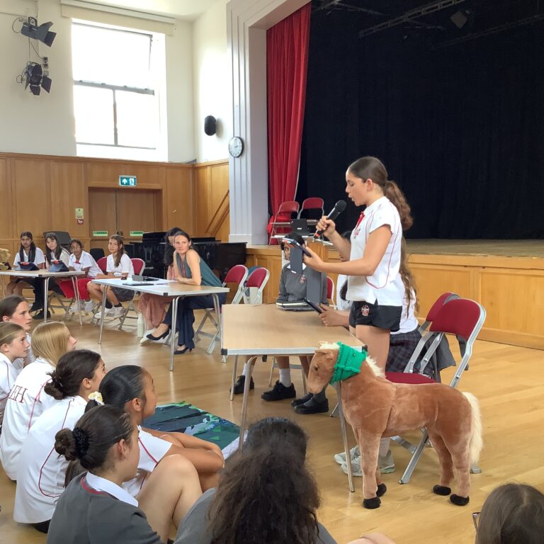 Student presenter speaks into a microphone at a desk in a school assembly, addressing seated classmates in white uniforms; a pony costume is in the foreground.