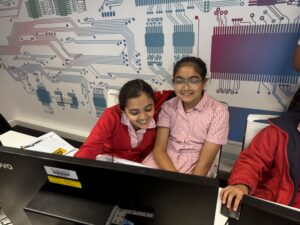 Two schoolgirls in red and striped uniforms sit at a desk with laptops, smiling, in front of a blue-purple circuit-wall mural in a classroom setting.