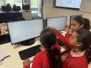 Group of students in red uniforms gathered around a desk, looking at computer screens displaying code in a bright classroom.
