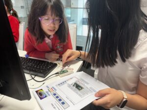 Young girl with pink glasses and a red cardigan concentrates on wiring a circuit on a white lab table, with a laptop nearby and an instruction sheet on the desk.
