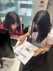 Two girls sit at a desk working on an electronics project, with a laptop, a small breadboard, and a printed diagram or worksheet in front of them at an office or classroom.