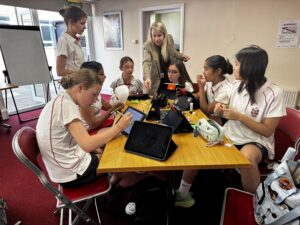 Teacher guiding a group of girls around a table with tablets and craft materials in a classroom.
