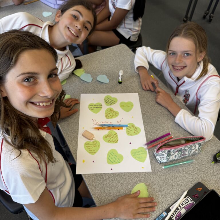 Group of students in school uniforms at a table making a heart-themed science project with green paper cutouts and markers.