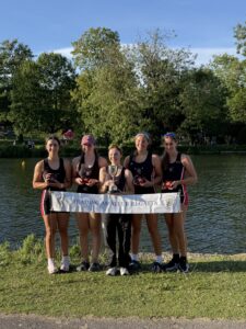 Five young rowers in black uniforms stand on a riverbank holding medals and a trophy, with a Reading Amateur Regatta banner in front of them.