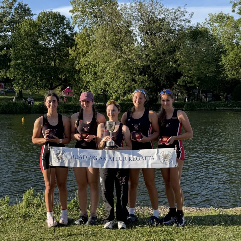 Five young rowers in black uniforms stand on a riverbank holding medals and a trophy, with a Reading Amateur Regatta banner in front of them.
