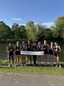 Group of young female rowers in black uniforms posing with trophies beside a lake, a park and trees in the background. Banner reads 'Reading Amateur Regatta'.