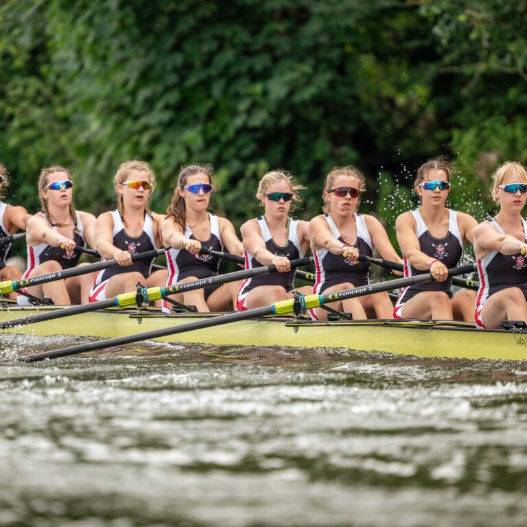 Eight female rowers in matching uniforms row a yellow racing shell on a river, splashing water as they propel forward with synchronized oars.