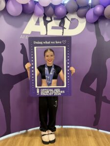Young dancer posing with a purple photo frame that reads 'Doing what we love' at a purple event backdrop with balloons and silhouettes; medal around neck.