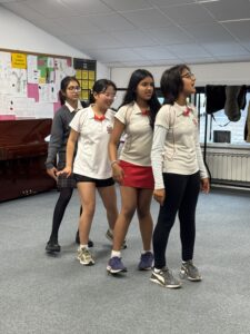 Four students in white polo shirts with red trim walk in a line across a classroom.